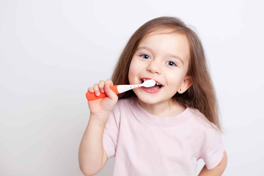 Mädchen im rosa Shirt putzt sich die Zähne mit einer Handzahnbürste und schaut dabei in die Kamera.